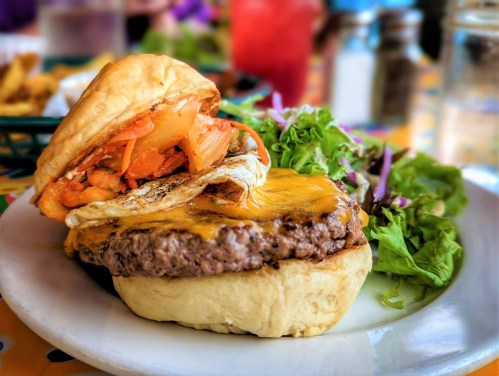 A juicy cheeseburger topped with grilled onions and a fried egg, served with a side salad on a colorful table.