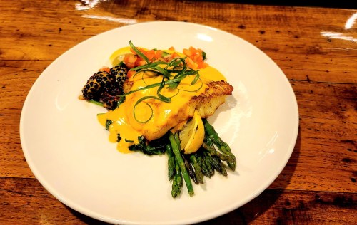 A plated dish featuring fish with a creamy sauce, asparagus, tomatoes, and a side of blackened vegetables on a wooden table.