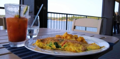 A plate with an omelette and diced potatoes, alongside a drink, with a scenic view of water in the background.