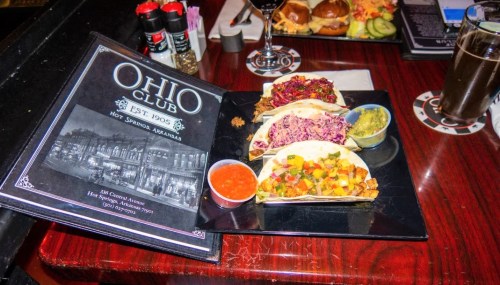 A plate of three tacos with colorful fillings, accompanied by salsa and guacamole, next to a menu from Ohio Club.