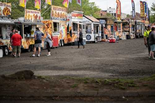 A row of food trucks with colorful banners, people ordering food in a gravel lot on a cloudy day.