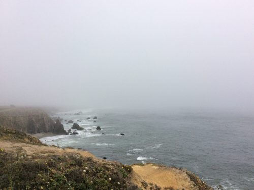 Foggy coastal scene with rocky cliffs and waves crashing against the shore, creating a serene, misty atmosphere.