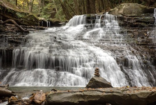 A serene waterfall cascades over rocky ledges, with a small stone stack in the foreground amidst autumn foliage.