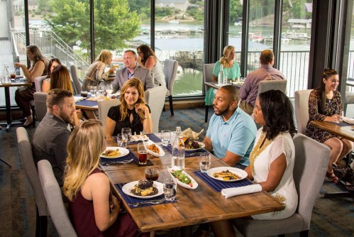 A group of people dining at a restaurant with large windows overlooking a lake, enjoying meals and conversation.