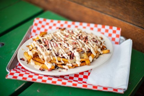 A plate of loaded fries topped with sauce and chopped toppings, served on a checkered paper with a napkin.