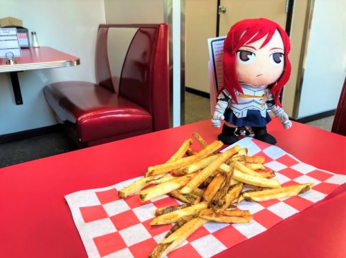 A plush doll with red hair stands beside a pile of golden fries on a red-checkered paper in a diner setting.