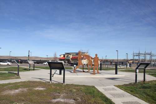 A metal sculpture of two figures in a park, with informational signs and a restaurant in the background under a clear sky.