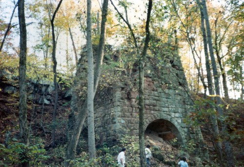 Ruins of a stone structure surrounded by trees in a forest during autumn, with two people exploring nearby.
