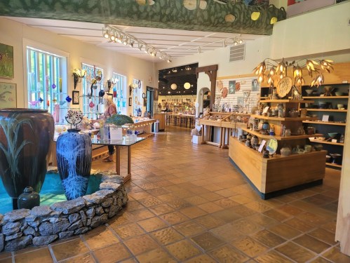 A bright art gallery featuring pottery, sculptures, and decorative items, with a stone fountain in the foreground.