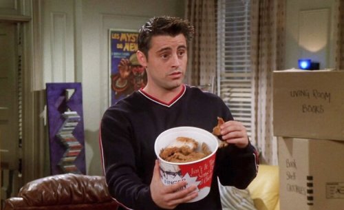A man holds a bucket of fried chicken, looking surprised, in a cozy living room with moving boxes around.