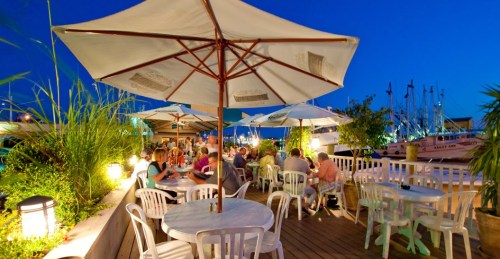 Outdoor dining area with tables and umbrellas, surrounded by greenery and boats, under a twilight sky.
