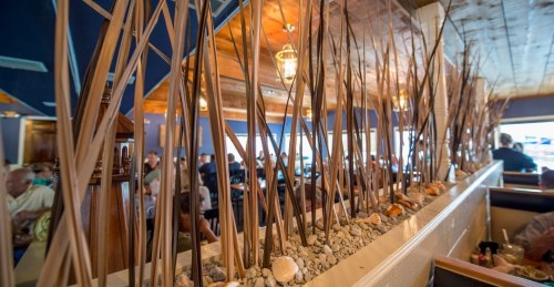 Interior of a restaurant featuring tall decorative grasses and pebbles, with diners visible in the background.