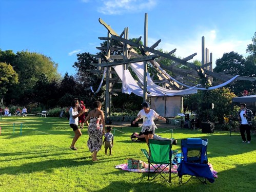 A lively outdoor gathering with people dancing on grass, colorful chairs, and a unique wooden structure in the background.