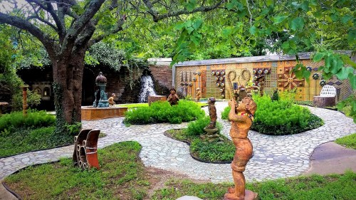 A serene garden featuring sculptures, a stone path, lush greenery, and a decorative wall with artistic designs.