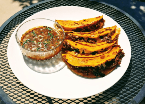 A plate of crispy tacos filled with meat, served with a side of rich sauce and garnished with herbs.