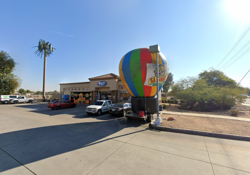 A colorful hot air balloon stands outside a store, with parked cars and palm trees in the background under a clear sky.
