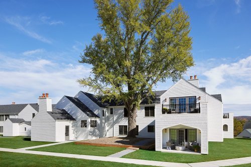 A modern white house with a large tree in the front yard, surrounded by green grass and a clear blue sky.