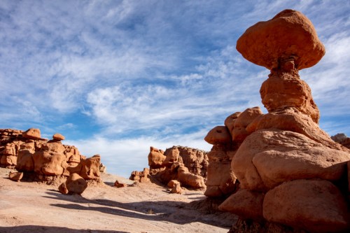 A unique rock formation stands tall in a desert landscape under a blue sky with scattered clouds.