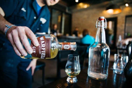 A person pours gin from a bottle into a glass at a bar, with a large water bottle in the background.