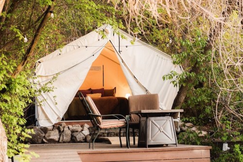 A cozy glamping tent nestled among trees, featuring a bed inside and outdoor seating on a wooden deck.