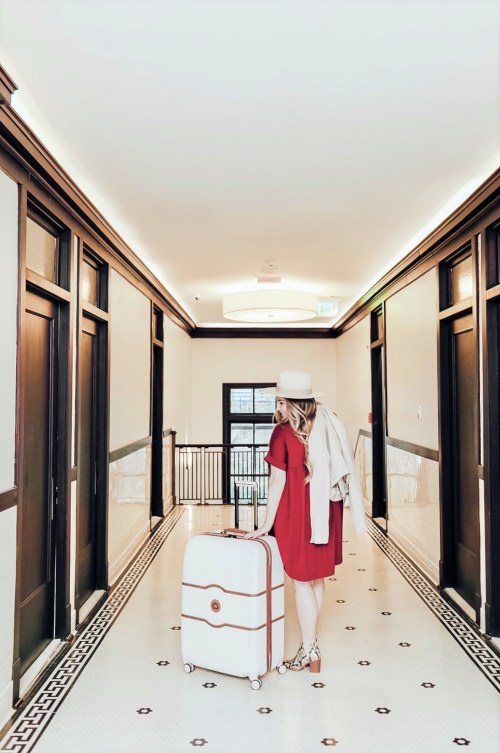 A woman in a red dress and hat stands in a stylish hotel hallway, pulling a suitcase behind her.