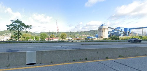 A view of a highway with a tree, an American flag, and industrial buildings in the background under a blue sky.