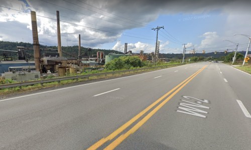 A view of a road alongside an industrial area with smokestacks and power lines under a cloudy sky.