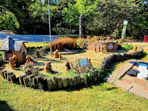 A whimsical garden featuring clay figures, flowers, and decorative elements, surrounded by a wooden fence and greenery.