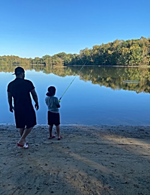 A person and a child fishing by a calm lake surrounded by trees on a clear day.