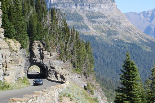 A car drives through a rocky tunnel on a scenic mountain road, surrounded by lush trees and towering cliffs.