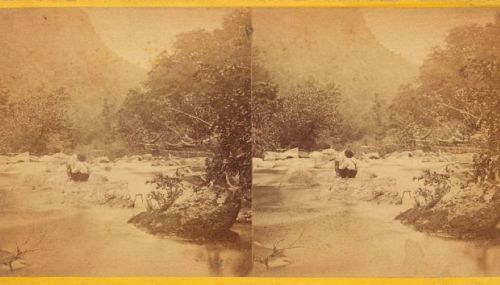 A vintage photograph of a river scene with a person standing among rocks and trees, captured in sepia tones.