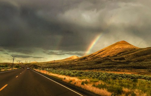 A scenic road leads towards mountains under a cloudy sky, with a vibrant rainbow arching over the landscape.