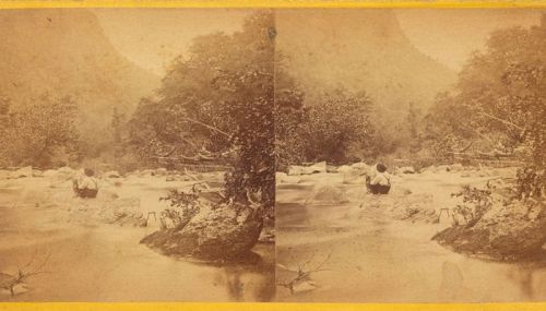 A vintage photograph of a river scene with a person standing by the water, surrounded by trees and rocky terrain.