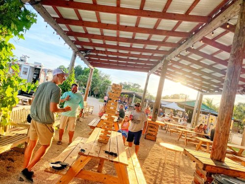 A group of people enjoying a sunny outdoor space, playing Jenga and socializing under a wooden structure.