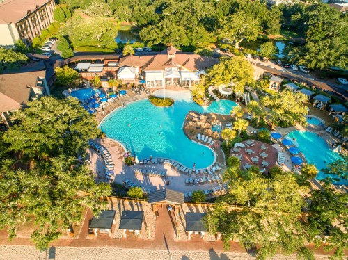 Aerial view of a resort pool area surrounded by lush greenery and lounge chairs, with a central building and water features.