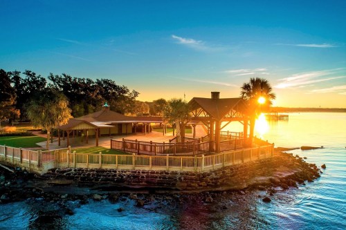 A scenic waterfront view at sunset, featuring a gazebo and palm trees along the shore.