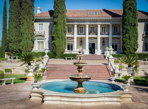 A grand mansion with a fountain in front, surrounded by lush greenery and tall cypress trees under a clear blue sky.