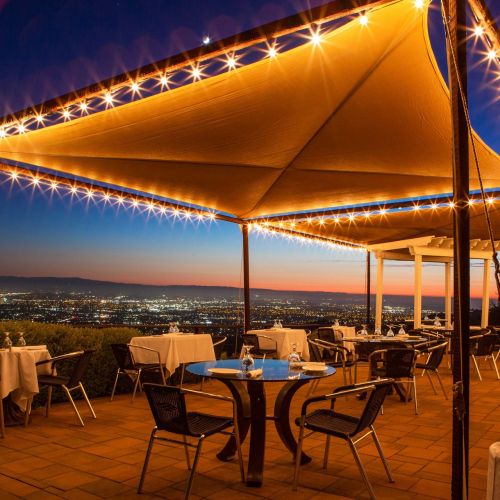 A beautifully lit outdoor dining area with tables, chairs, and a starry sky view over a cityscape at dusk.