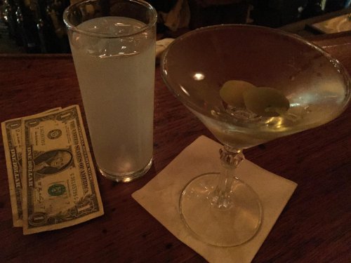 A glass of clear liquid, a martini with olives, and a dollar bill on a bar counter.