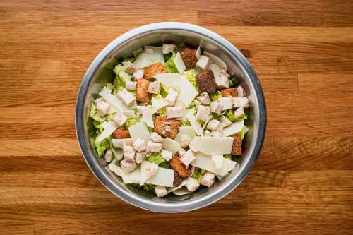 A bowl of salad with lettuce, diced chicken, croutons, and shaved cheese on a wooden surface.
