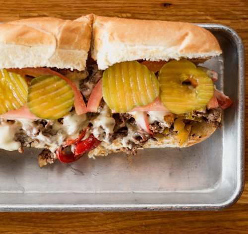 A loaded sandwich with sliced pickles, tomatoes, and melted cheese on a toasted hoagie roll, served on a metal tray.