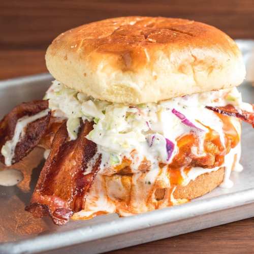 A hearty sandwich with crispy bacon, coleslaw, and a saucy filling, served on a toasted bun.