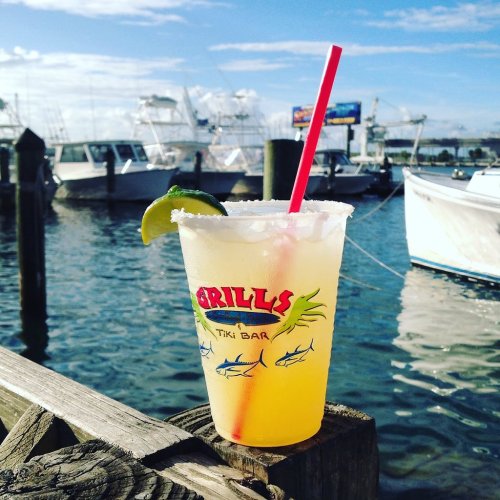A refreshing cocktail with a lime wedge sits on a wooden dock, with boats and a blue sky in the background.