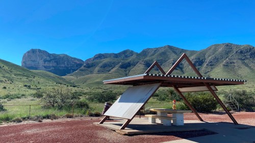A modern shelter with a picnic table in a scenic landscape of green hills and a prominent rock formation under a clear blue sky.