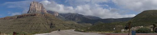 A panoramic view of a mountainous landscape with a prominent rock formation and a winding road under a partly cloudy sky.