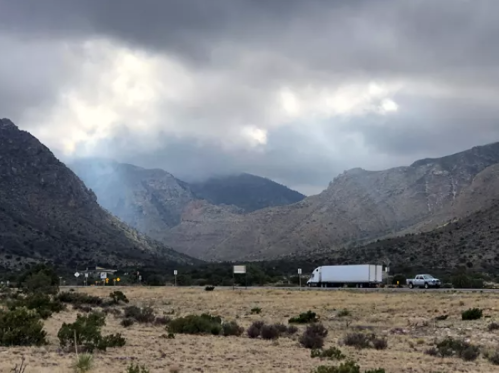 A truck and a car parked near a highway, surrounded by mountains under a cloudy sky. Smoke rises in the distance.