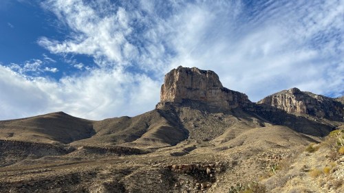 A rugged mountain peak rises above a dry landscape under a blue sky with scattered clouds.