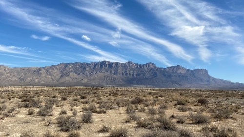 A vast desert landscape with sparse vegetation and a mountain range under a blue sky with wispy clouds.