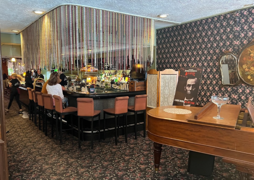 A stylish bar with a vintage decor, featuring a long counter, colorful lights, and a piano in the foreground.