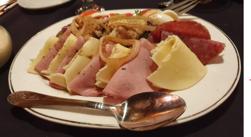 A platter of assorted cold cuts, cheeses, and garnishes, served with a spoon on a black tablecloth.
